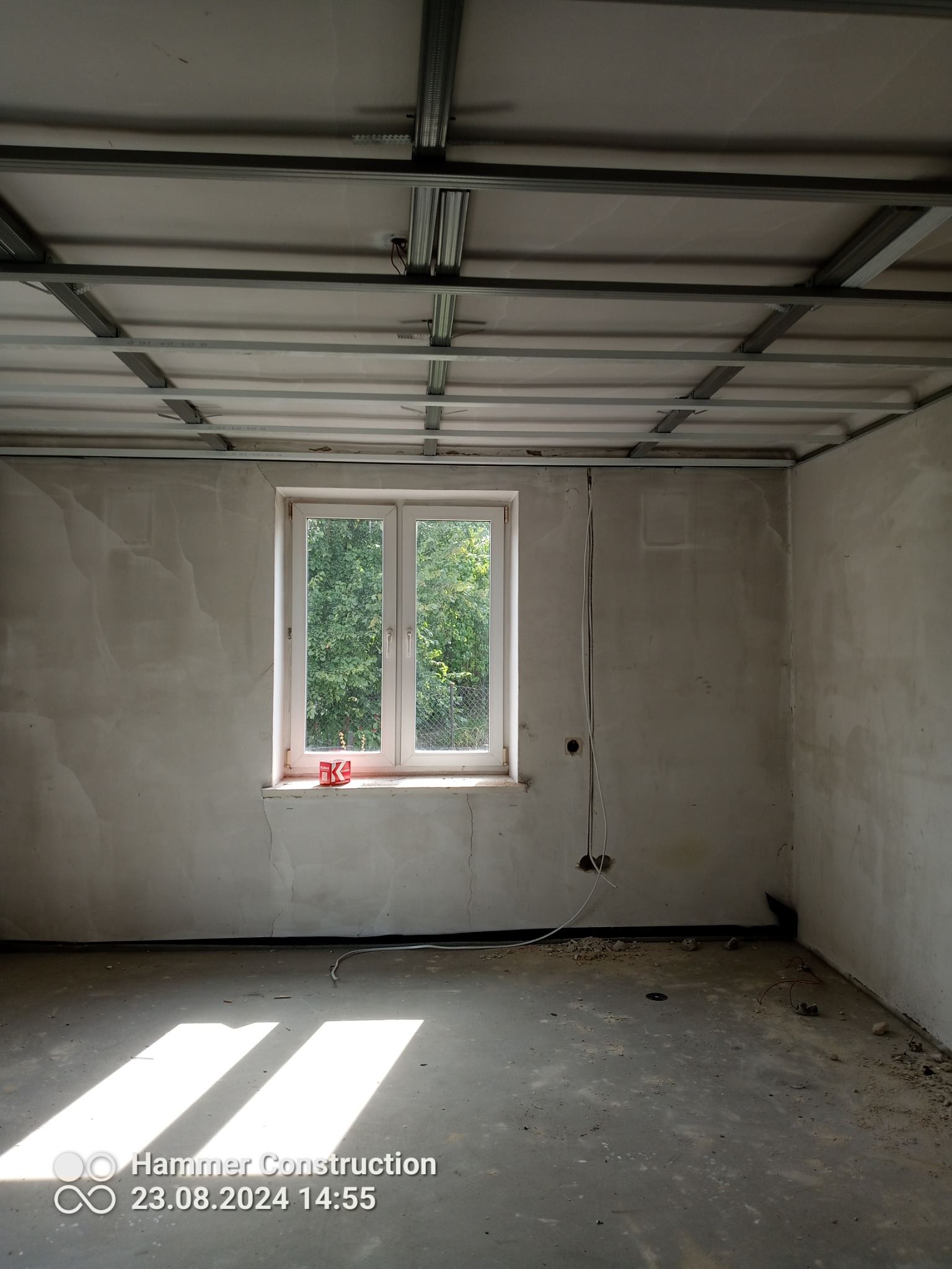 Wnętrze w trakcie remontu: surowe ściany z widocznymi śladami gładzi, metalowa konstrukcja podwieszanego sufitu, okno z widokiem na zieleń, wystające kable elektryczne.