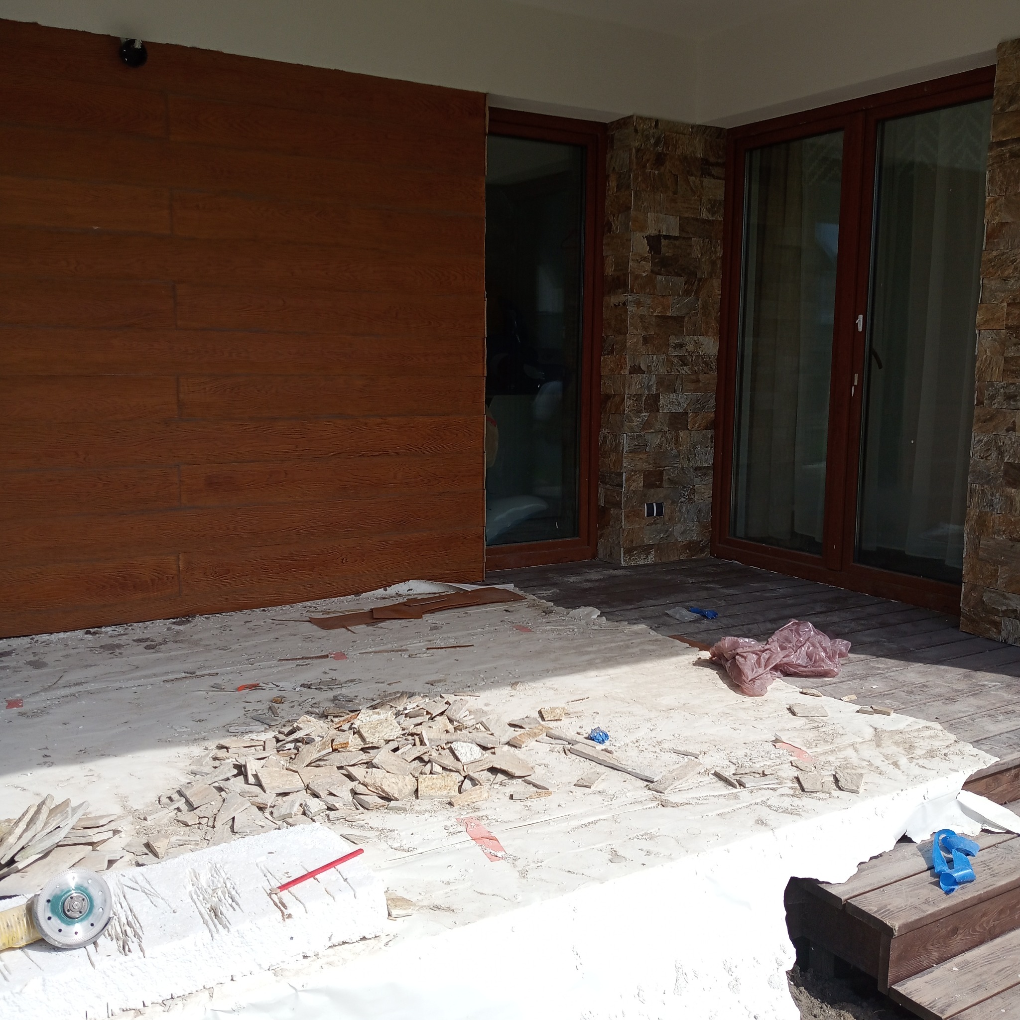 Fragment tarasu z drewnianą podłogą w trakcie prac remontowych, z widocznymi płytkami elewacyjnymi, narzędziami (szlifierka kątowa) i folią ochronną, na tle ściany z okładziną drewnianą i kamienną.