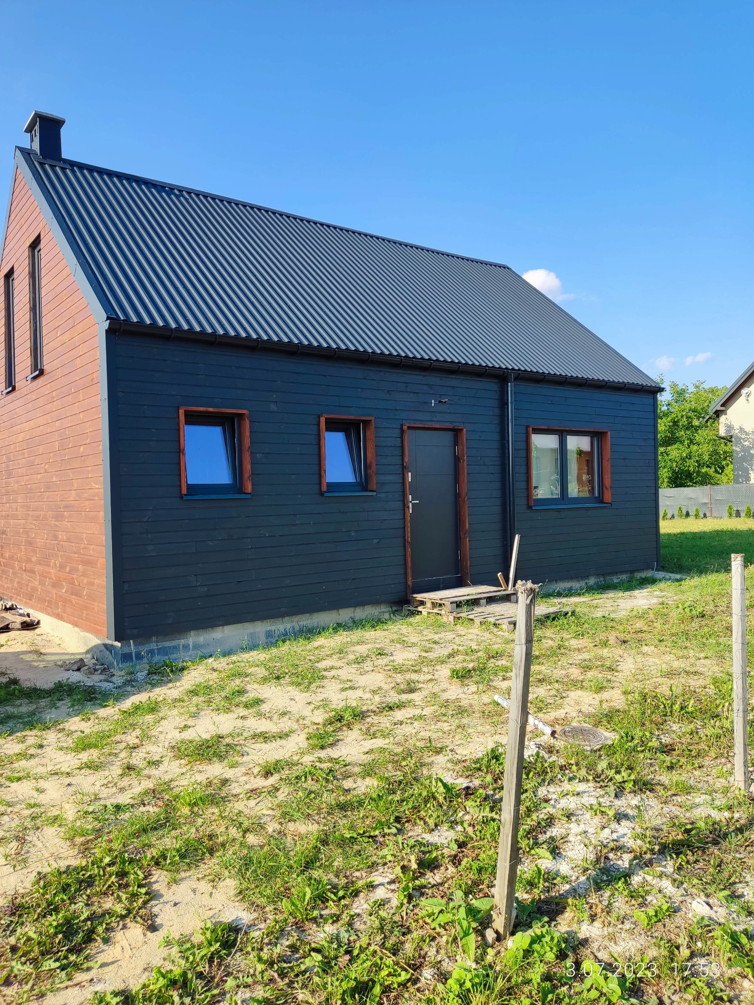 Nowoczesny dom z czarną elewacją i grafitowym dachem z blachy trapezowej, drewniane wykończenia okien i fragment brązowej ściany szczytowej, widoczne palety przed wejściem.