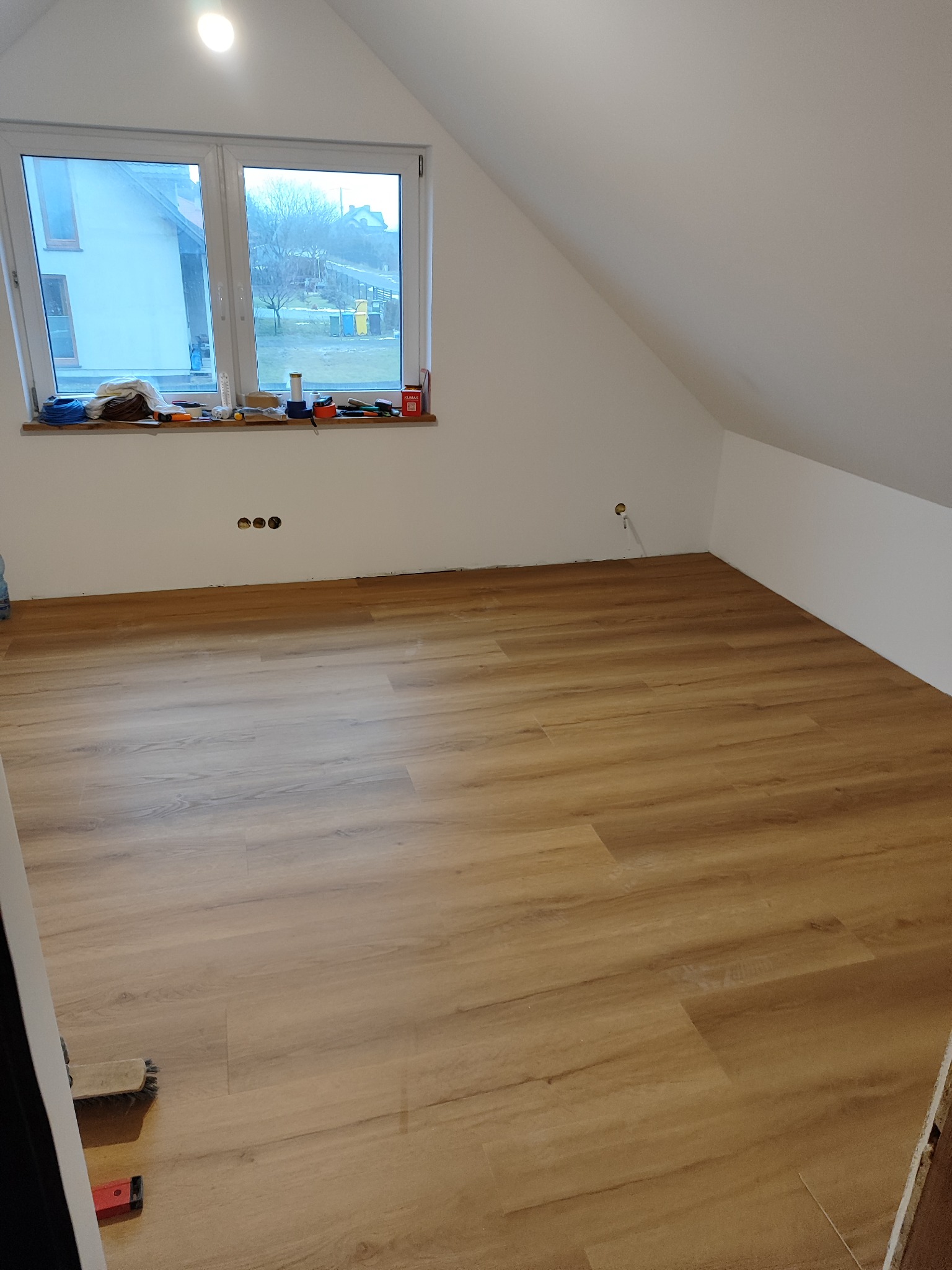 Nowo położona podłoga z paneli w jasnym odcieniu drewna w pokoju na poddaszu, widoczne narzędzia na parapecie okna.