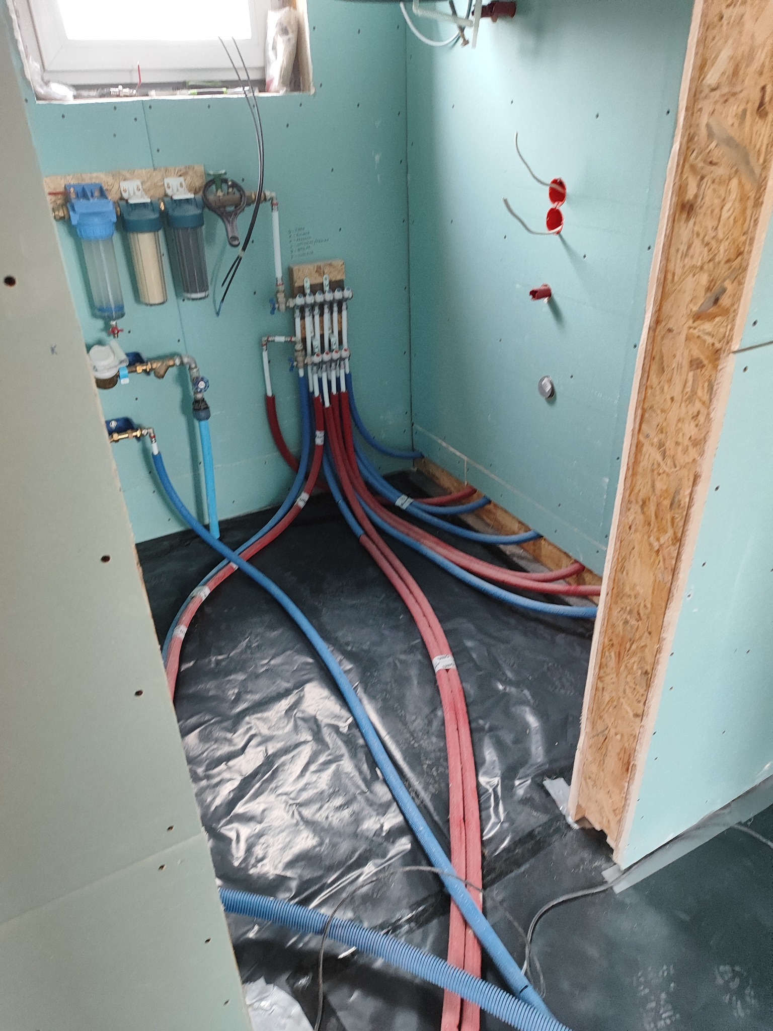 Instalacja wodna w trakcie montażu w pomieszczeniu z płytami gipsowo-kartonowymi, widoczne filtry wody, rozdzielacz z czerwonymi i niebieskimi rurami oraz wystające przewody elektryczne ze ściany.