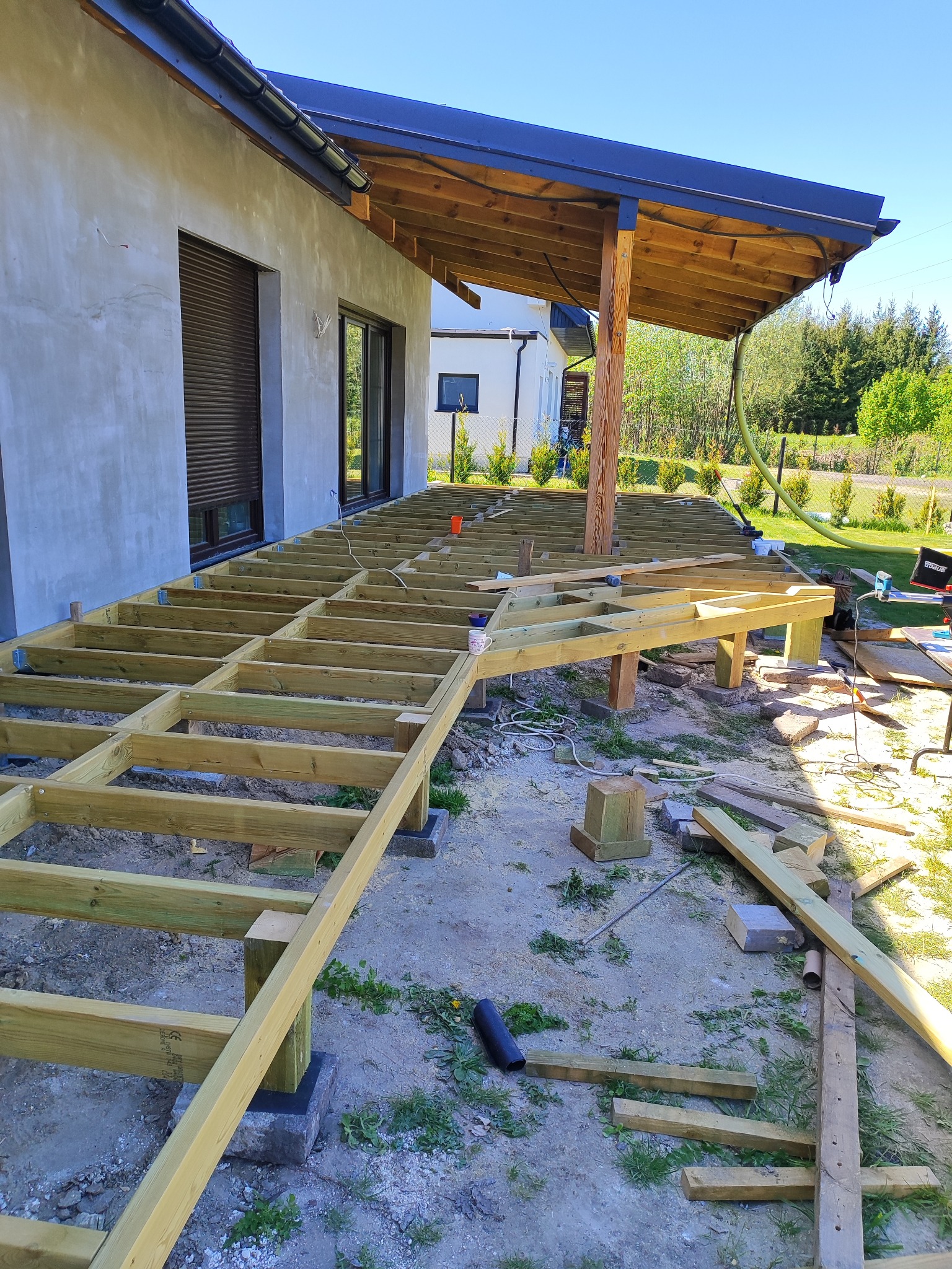 Konstrukcja nośna tarasu z impregnowanego drewna, widoczna w trakcie montażu przy budynku z szarą elewacją i drewnianym zadaszeniem, narzędzia i materiały budowlane wokół.