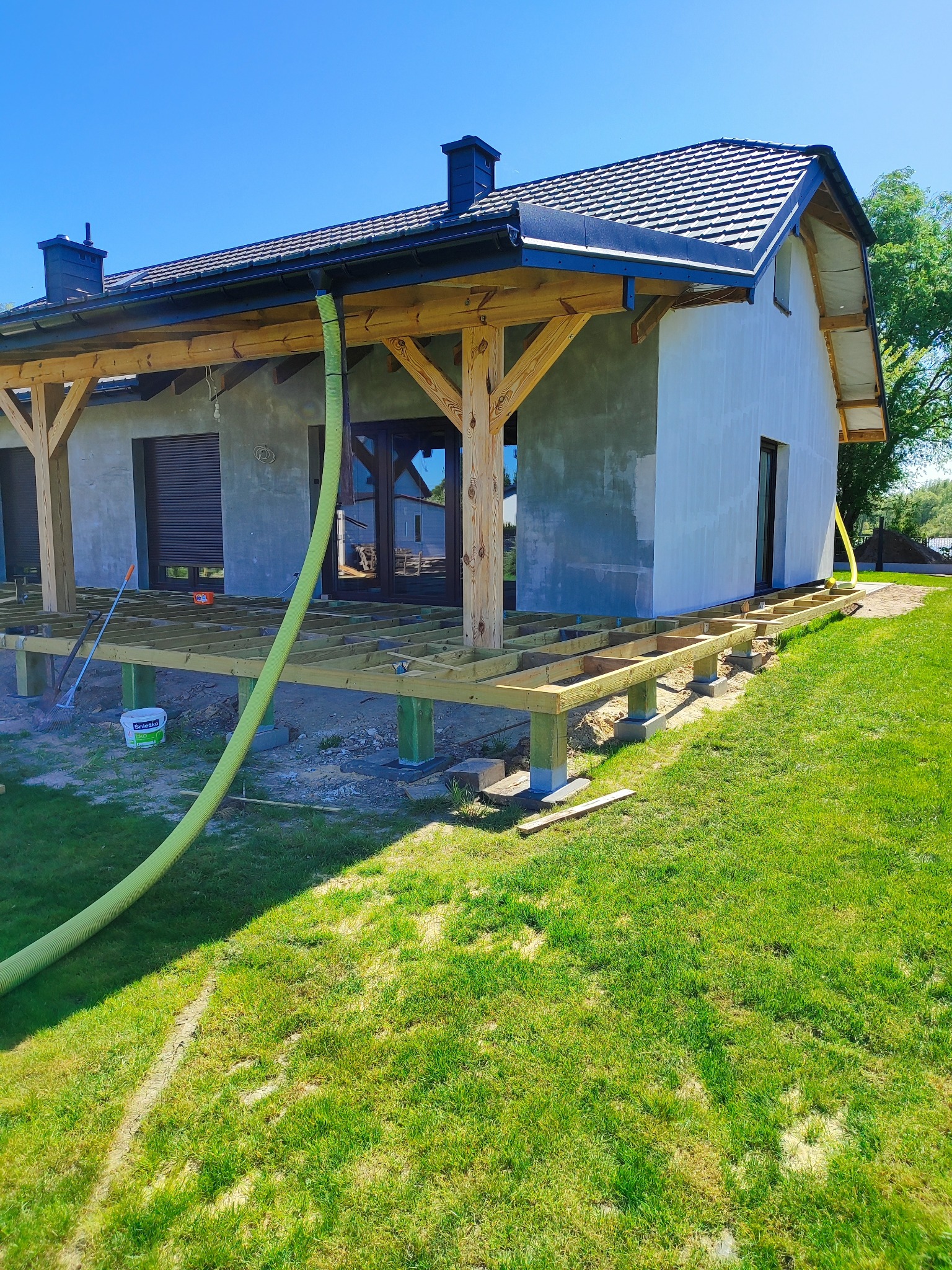 Konstrukcja drewnianego tarasu w trakcie budowy przy domu z szarą elewacją, widoczna zielona rura wentylacyjna i podłoże z trawy.