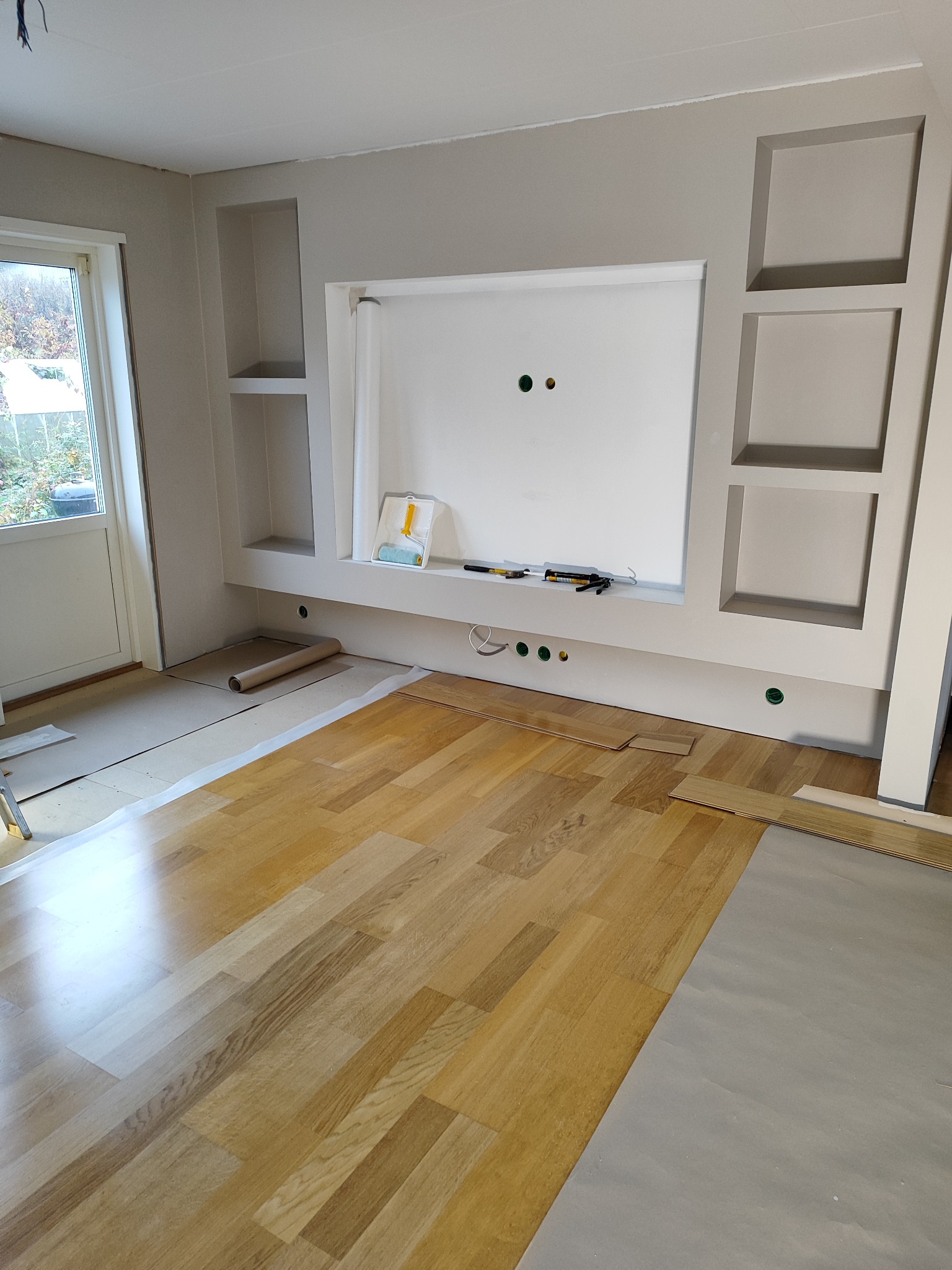 Wnętrze pokoju w trakcie remontu: częściowo położona drewniana podłoga, zabezpieczona folią, ściana z wnękami pomalowana na szaro, widoczne narzędzia i materiały.