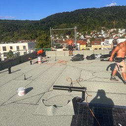 Dachyix Sp.Z.O.O - Prace na dachu płaskim z zieloną papą termozgrzewalną BauderTEC KSA DUO 35, ekipa dekarzy w trakcie montażu, widoczne narzędzia i materiały, w tle zabudowa miejska i zalesione wzgórze.