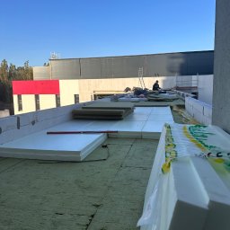 Dachyix Sp.Z.O.O - Ocieplenie dachu: styropian i wełna mineralna na zielonej membranie. W tle budynek z czerwoną ścianą i pracownik na drabinie. Poziomica na izolacji.