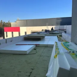 Ocieplenie dachu: styropian i wełna mineralna na zielonej membranie. W tle budynek z czerwoną ścianą i pracownik na drabinie. Poziomica na izolacji.