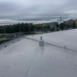 Jasnoszary dach płaski z widocznymi elementami wentylacyjnymi i uszczelniającymi na tle bloków mieszkalnych i pochmurnego nieba.