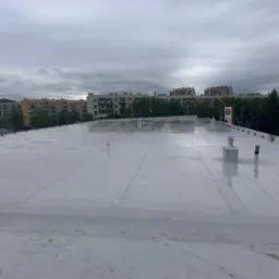 Biały, płaski dach po deszczu w Miękini, z widocznymi kominami wentylacyjnymi i budynkami w tle. Dach pokryty membraną PVC, świeżo po montażu.