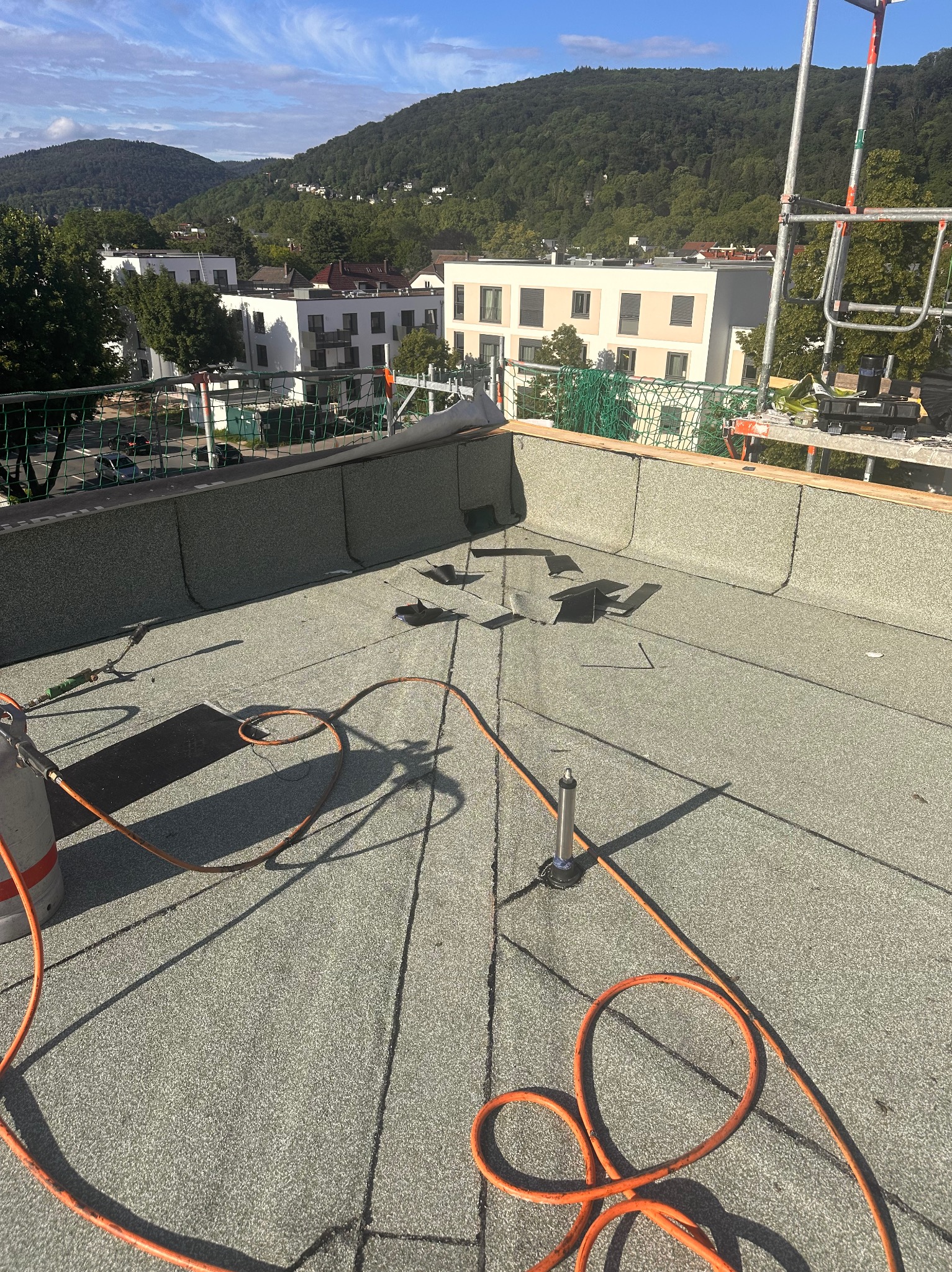 Nowe pokrycie papą termozgrzewalną na płaskim dachu budynku, widoczne narzędzia, palnik i butla gazowa, w tle zabudowa miejska i zalesione wzgórza.