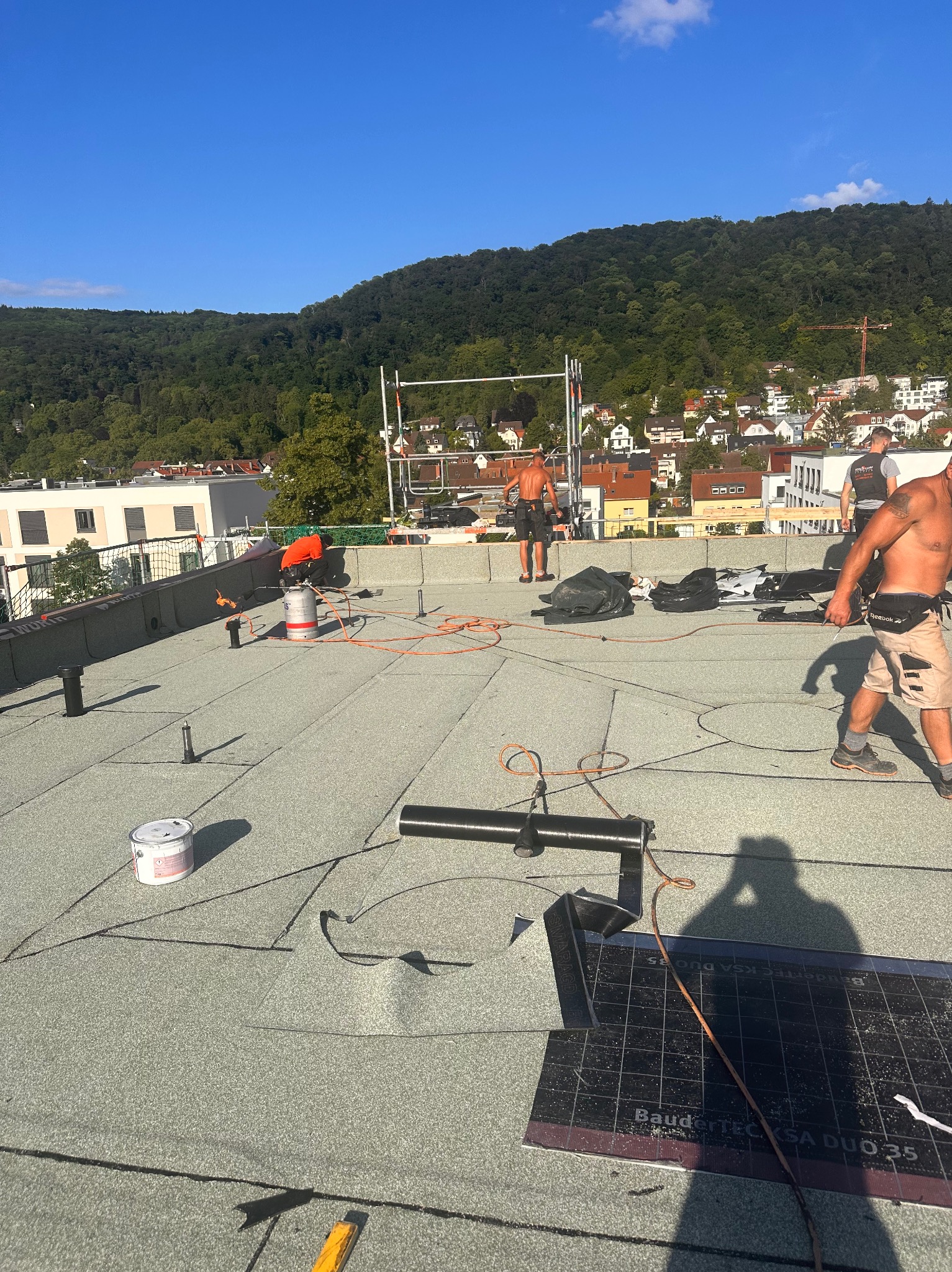Prace na dachu płaskim z zieloną papą termozgrzewalną BauderTEC KSA DUO 35, ekipa dekarzy w trakcie montażu, widoczne narzędzia i materiały, w tle zabudowa miejska i zalesione wzgórze.