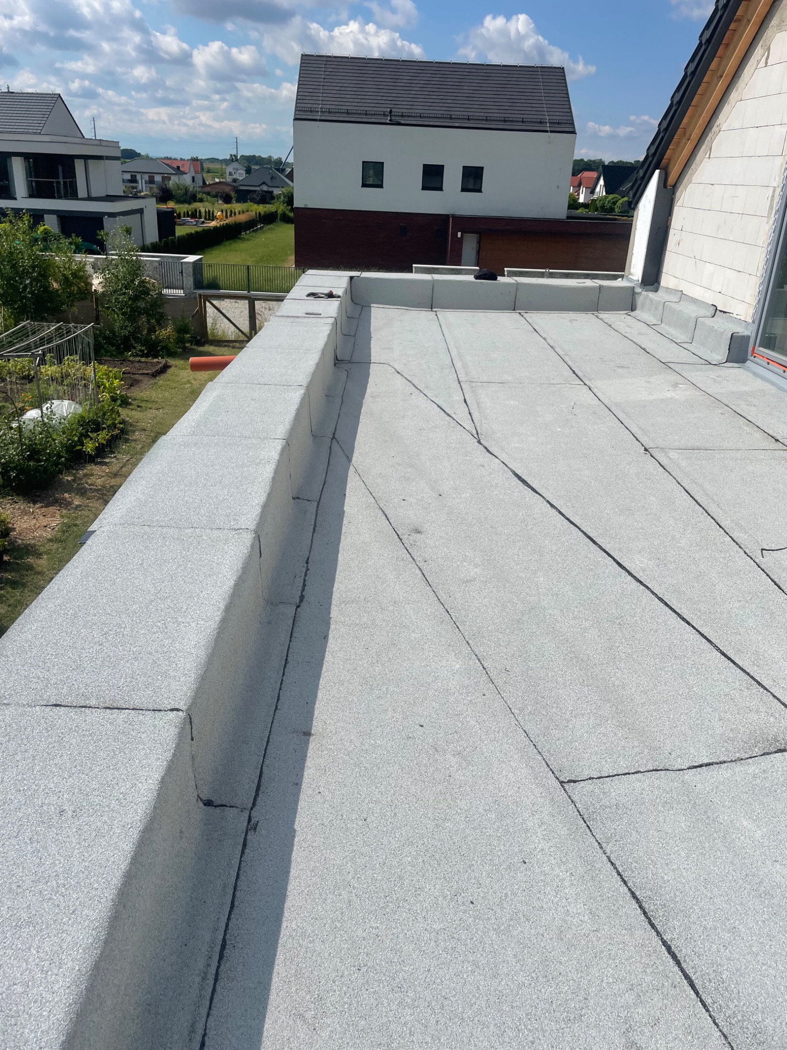 Szary, płaski dach pokryty papą termozgrzewalną, z widocznymi łączeniami i obróbkami. W tle zabudowa jednorodzinna i błękitne niebo.