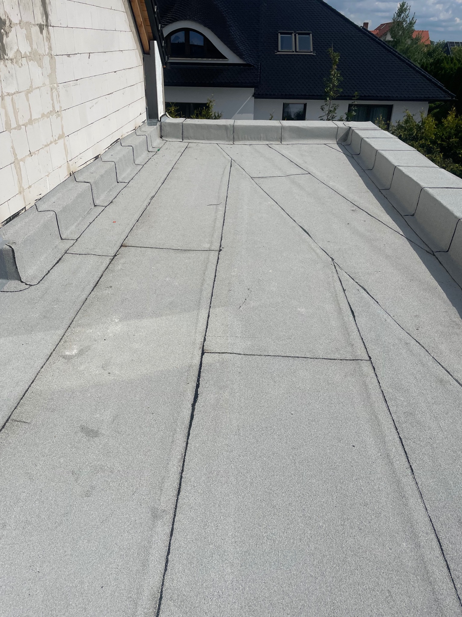 Szary dach pokryty papą termozgrzewalną, widok z góry, z widocznymi detalami łączeń i obróbek blacharskich. W tle budynek z czarnym dachem.