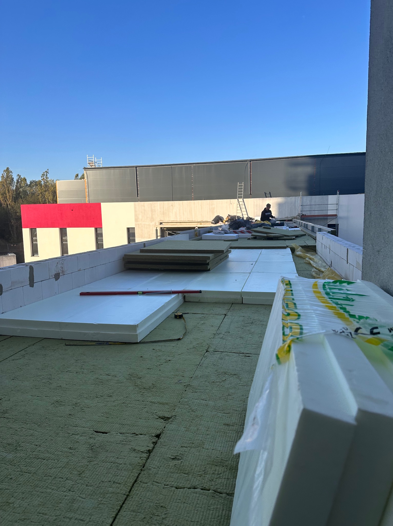 Ocieplenie dachu: styropian i wełna mineralna na zielonej membranie. W tle budynek z czerwoną ścianą i pracownik na drabinie. Poziomica na izolacji.