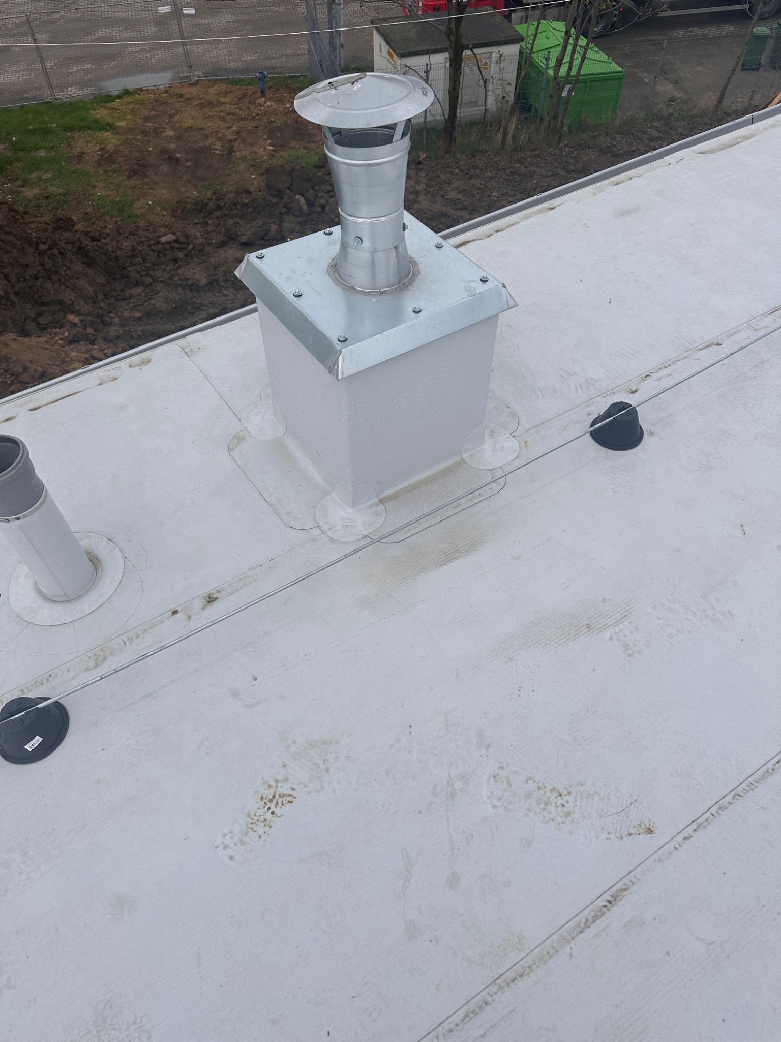 Szary, płaski dach z metalowym kominem wentylacyjnym i elementami mocującymi. Widoczne ślady zabrudzeń i zarysowania na powierzchni dachu.