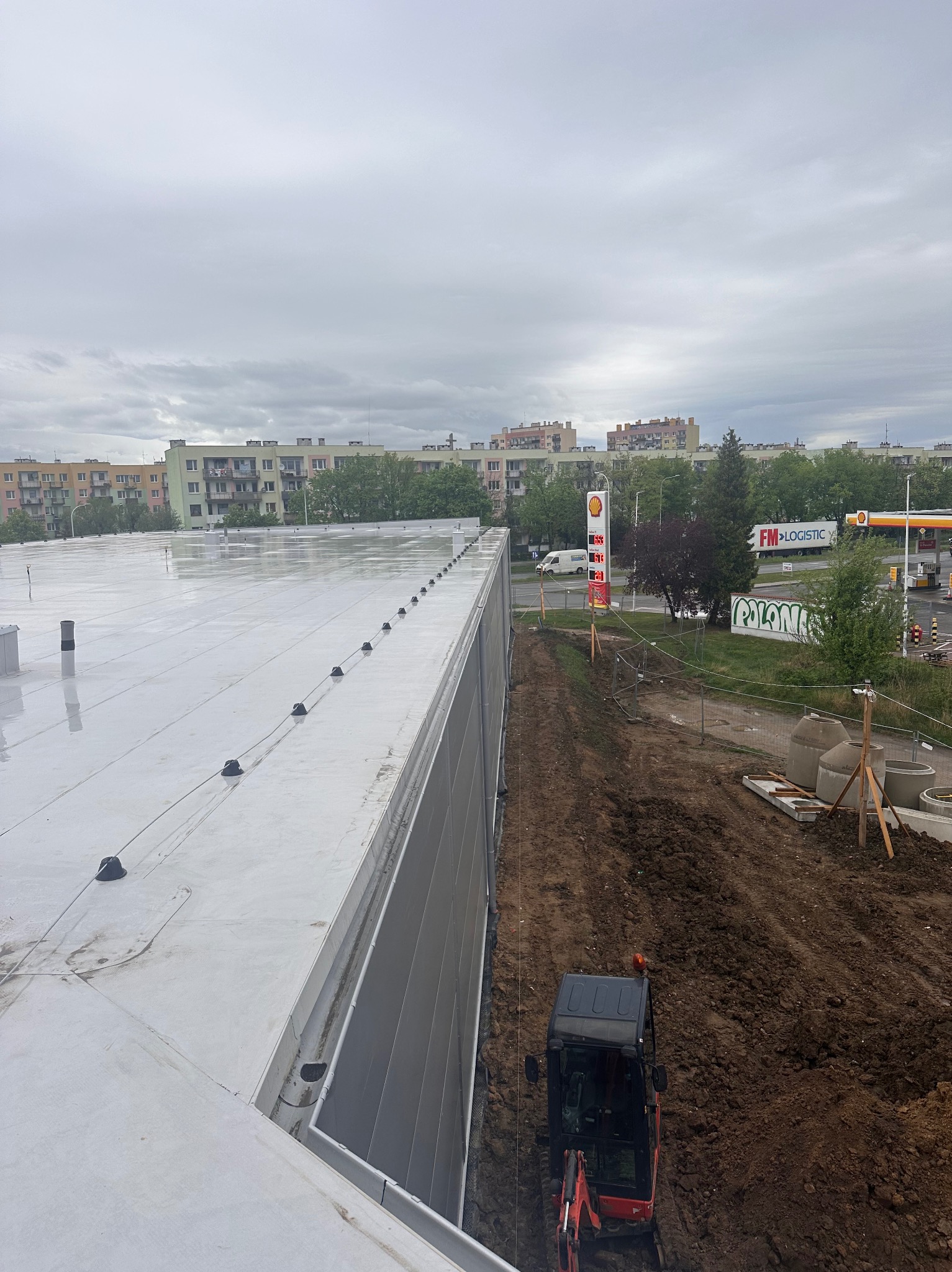 Jasnoszary, płaski dach budynku przemysłowego po deszczu, widok z góry. W tle zabudowa miejska i stacja benzynowa. Na dole mała koparka.