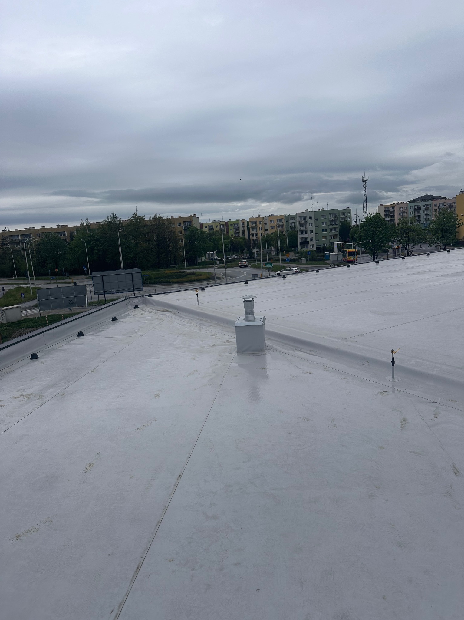 Jasnoszary dach płaski z widocznymi elementami wentylacyjnymi i uszczelniającymi na tle bloków mieszkalnych i pochmurnego nieba.