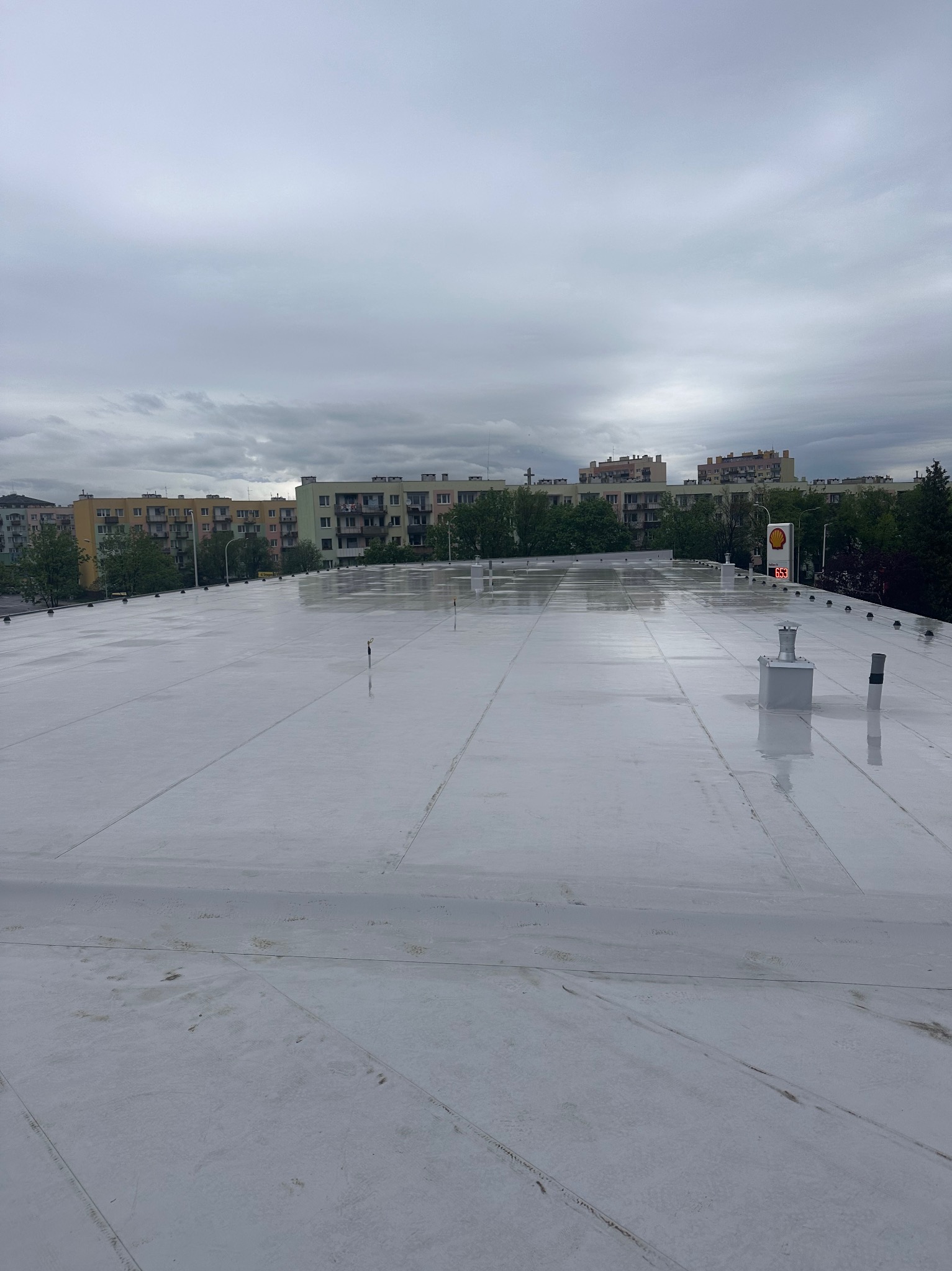 Biały, płaski dach po deszczu w Miękini, z widocznymi kominami wentylacyjnymi i budynkami w tle. Dach pokryty membraną PVC, świeżo po montażu.