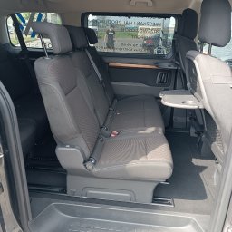AV PROFI SERWIS SPÓŁKA Z OGRANICZONĄ ODPOWIEDZIALNOŚCIĄ - Wnętrze busa pasażerskiego z trzema rzędami siedzeń, rozkładanymi stolikami i widokiem na szybę z napisem 'Przewozy Osobowe'.