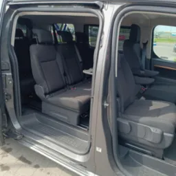 Szare auto typu van z otwartymi drzwiami suwanymi, ukazujące wnętrze z trzema rzędami siedzeń. Widoczne fotele z zagłówkami i pasami bezpieczeństwa, stolik rozkładany.