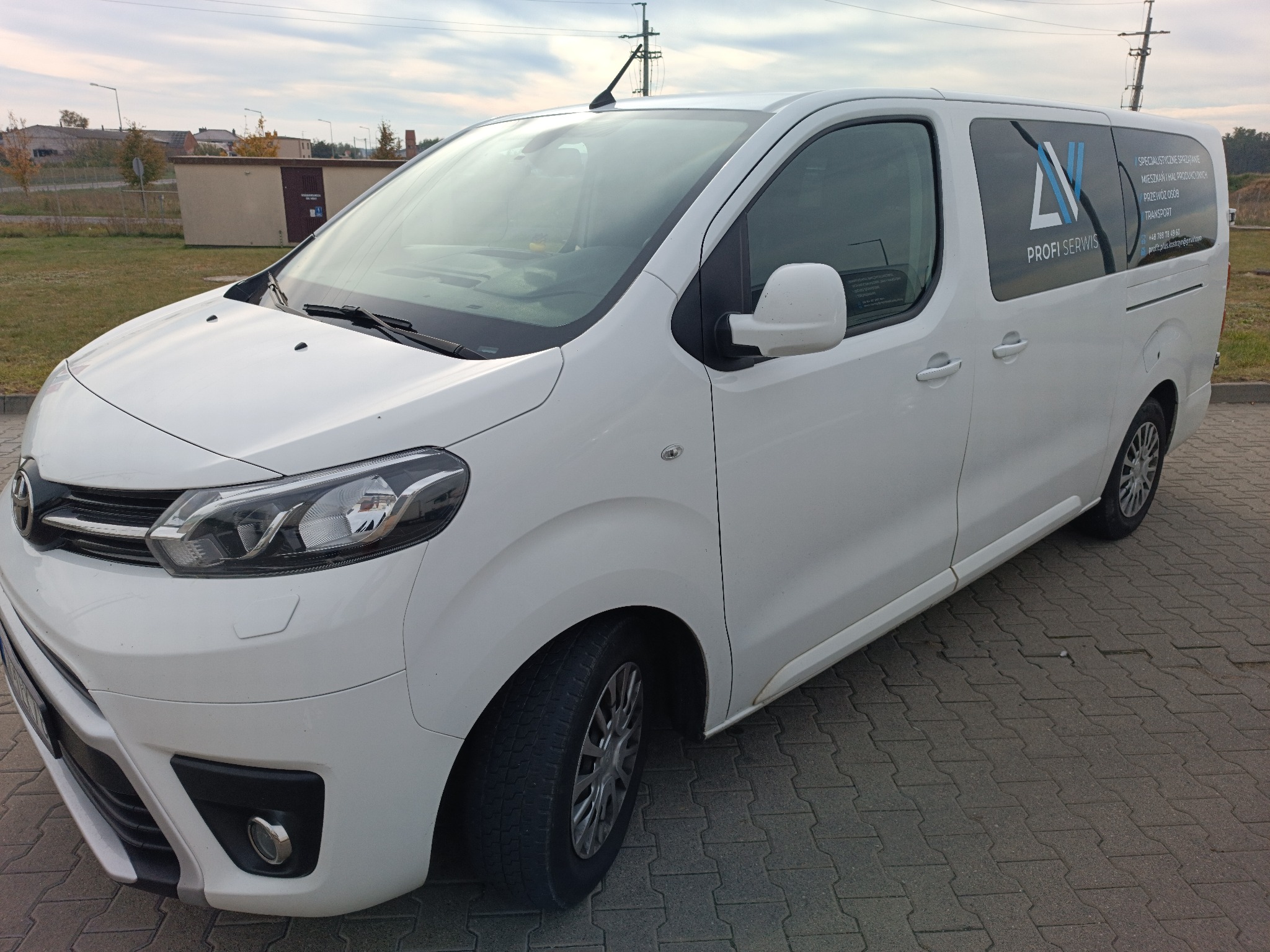 Biały bus z logo firmy i napisem 'Przewóz osób' na bocznej szybie, zaparkowany na kostce brukowej. Widoczne budynki w tle i pochmurne niebo.