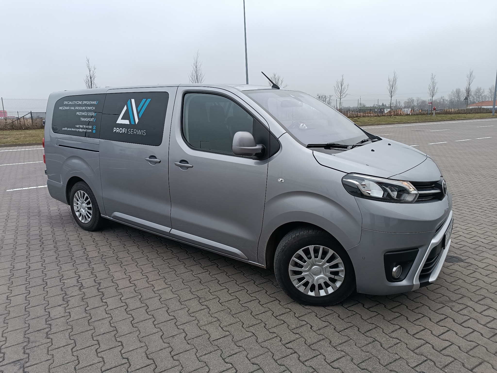 Szary van z logo 'Profi Serwis' na przyciemnianej szybie, zaparkowany na brukowanej powierzchni. Widoczne drzewa w tle i pochmurne niebo. Reklama usług sprzątania i transportu.
