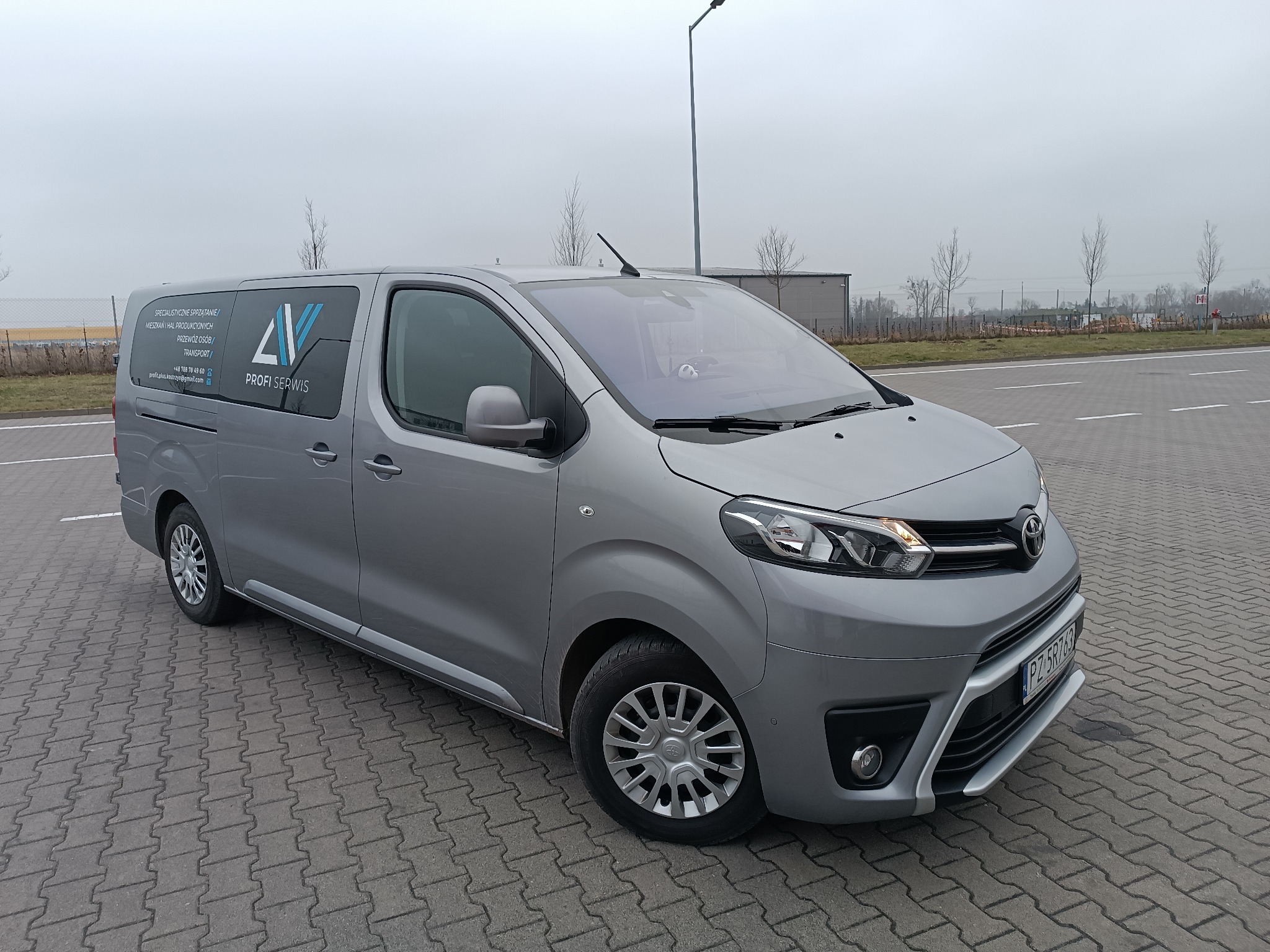 Szary van Toyota Proace Verso z logo firmy przewozowej na boku, zaparkowany na brukowanym placu. Na tablicy rejestracyjnej widoczny numer.