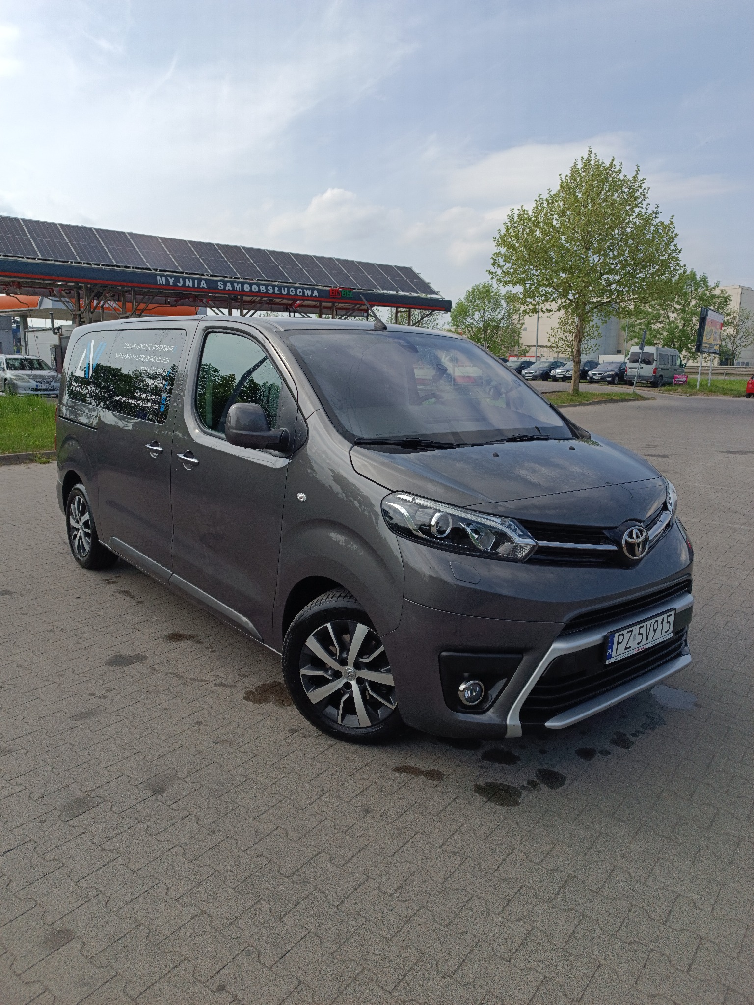 Szary van Toyota Proace Verso na kostce brukowej przed myjnią samoobsługową. Na szybie widoczne logo firmy sprzątającej. Tablica rejestracyjna PZ 5V915.