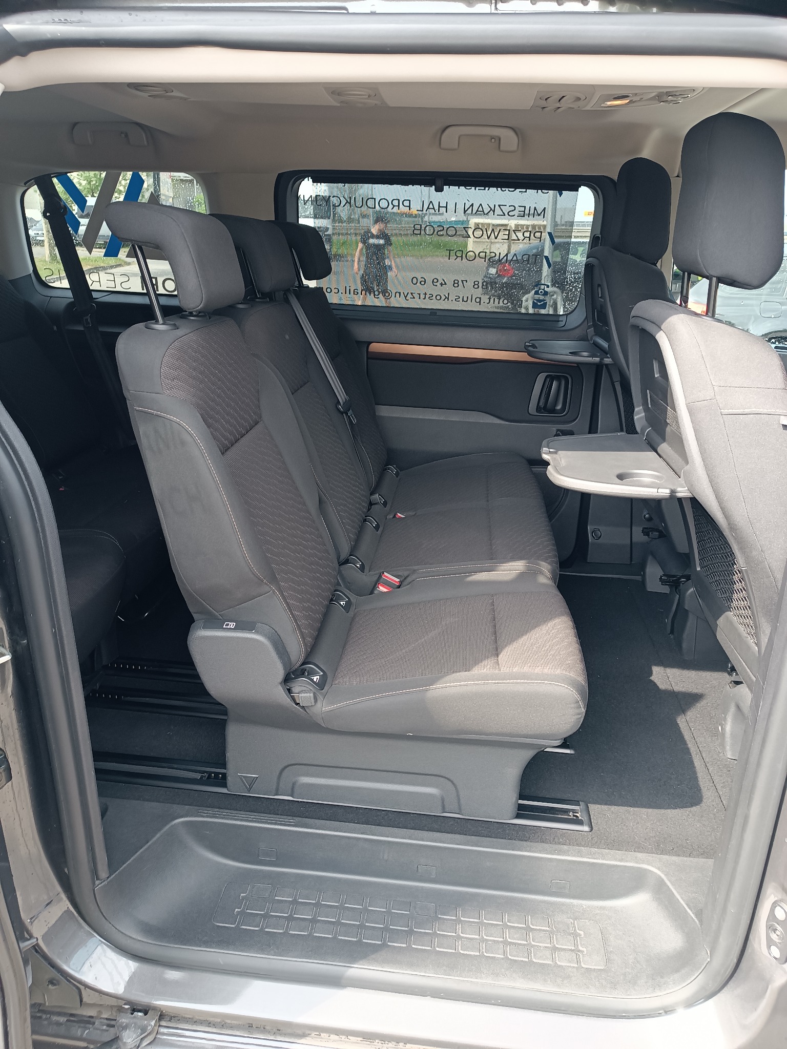 Wnętrze busa pasażerskiego z trzema rzędami siedzeń, rozkładanymi stolikami i widokiem na szybę z napisem 'Przewozy Osobowe'.