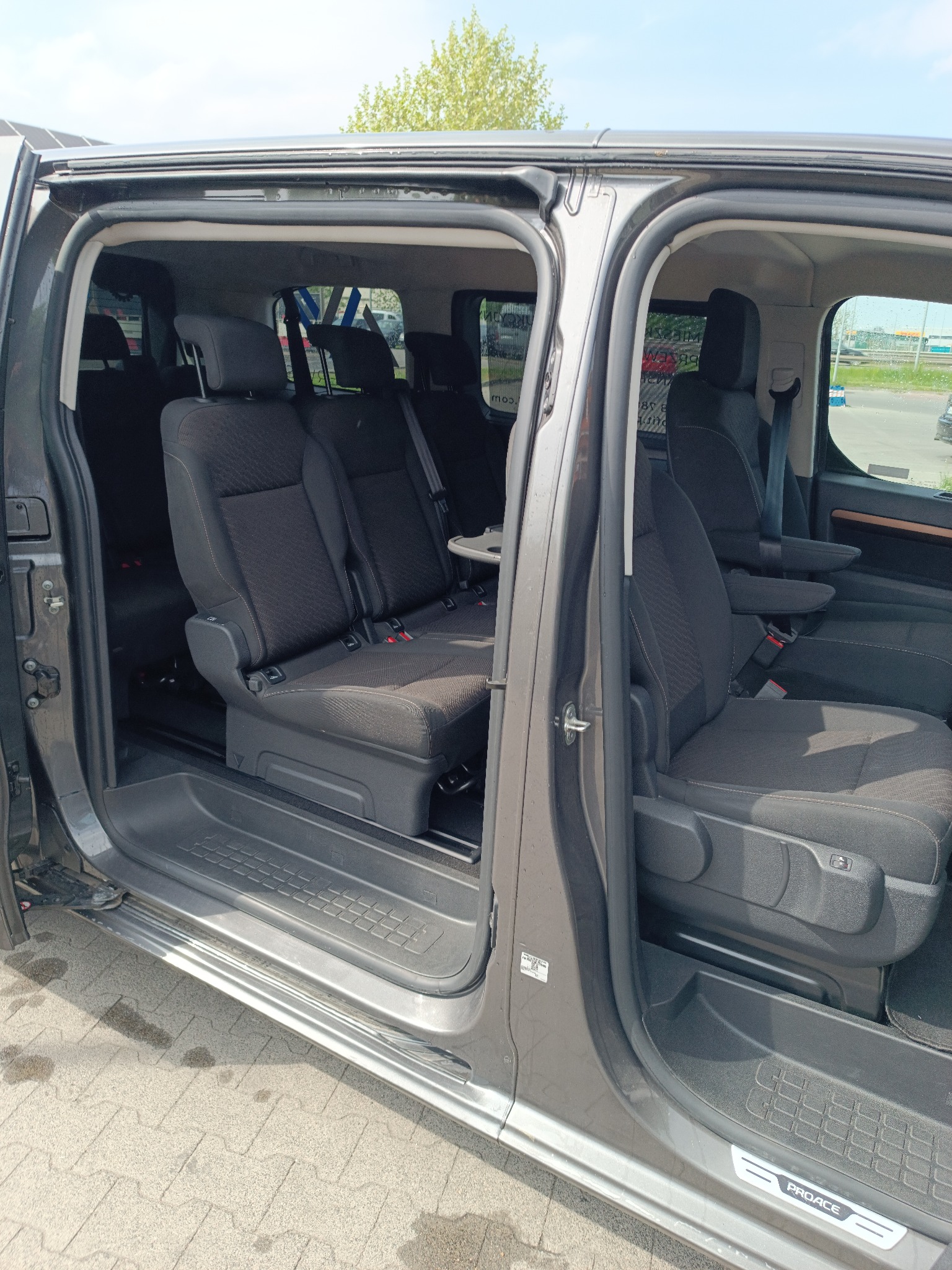 Szare auto typu van z otwartymi drzwiami suwanymi, ukazujące wnętrze z trzema rzędami siedzeń. Widoczne fotele z zagłówkami i pasami bezpieczeństwa, stolik rozkładany.