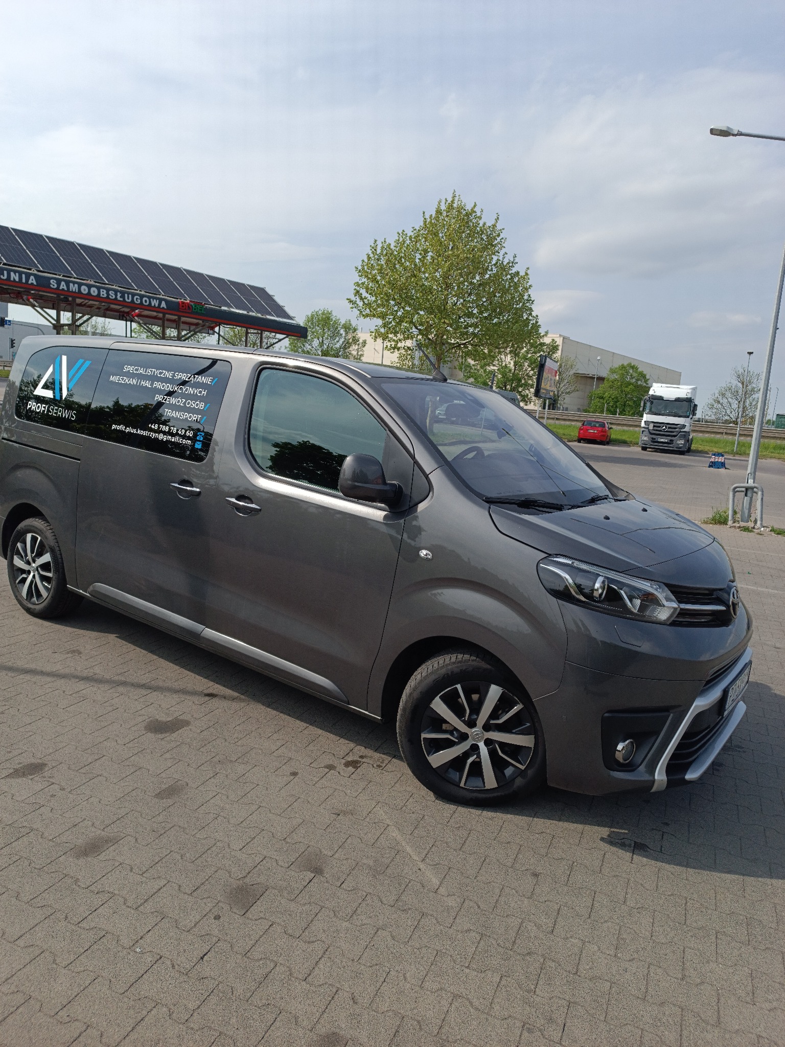 Szary van z logo firmy Profi Serwis na tle parkingu i myjni samochodowej. Na szybie informacje o sprzątaniu, transporcie i numer telefonu.