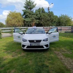 Biały samochód osobowy z otwartymi drzwiami, oznaczony jako taksówka, zaparkowany na trawniku przed ogrodzeniem.