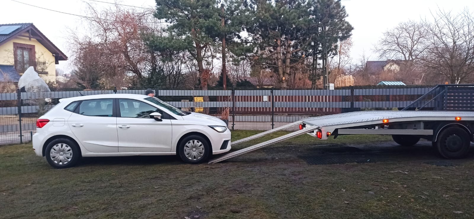 Biały samochód osobowy wjeżdża na najazd lawety na trawiastym terenie przed domem jednorodzinnym z szarym ogrodzeniem w tle.