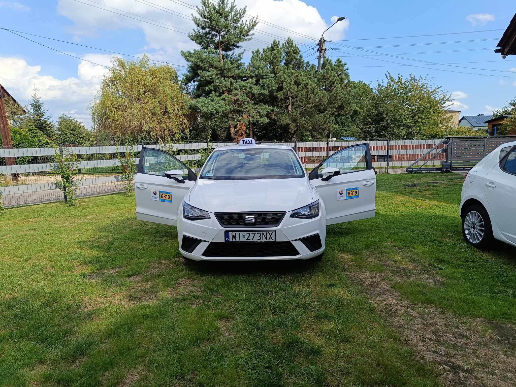 Biały samochód osobowy z otwartymi drzwiami, oznaczony jako taksówka, zaparkowany na trawniku przed ogrodzeniem.