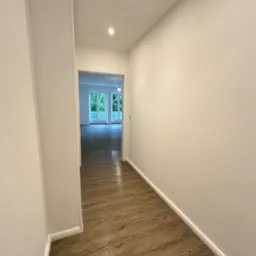 Wnętrze wykończonego korytarza z panelami podłogowymi imitującymi drewno i białymi ścianami, widok na przeszklone drzwi balkonowe w głębi