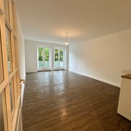 Usługi Ogólnobudowlane Oliwia Ochmańska - Jasne, przestronne wnętrze po remoncie z nową podłogą imitującą drewno, świeżo pomalowanymi ścianami i otwartymi, przeszklonymi drzwiami balkonowymi z widokiem na zieleń.