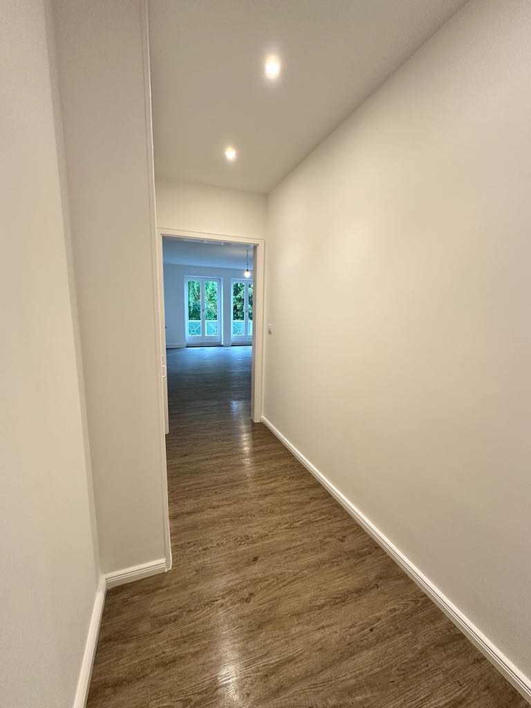 Wnętrze wykończonego korytarza z panelami podłogowymi imitującymi drewno i białymi ścianami, widok na przeszklone drzwi balkonowe w głębi