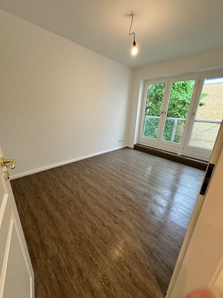 Wnętrze pokoju z drewnopodobną podłogą, białymi ścianami i dużymi oknami balkonowymi z widokiem na zieleń, widoczna żarówka na kablu zwisająca z sufitu.