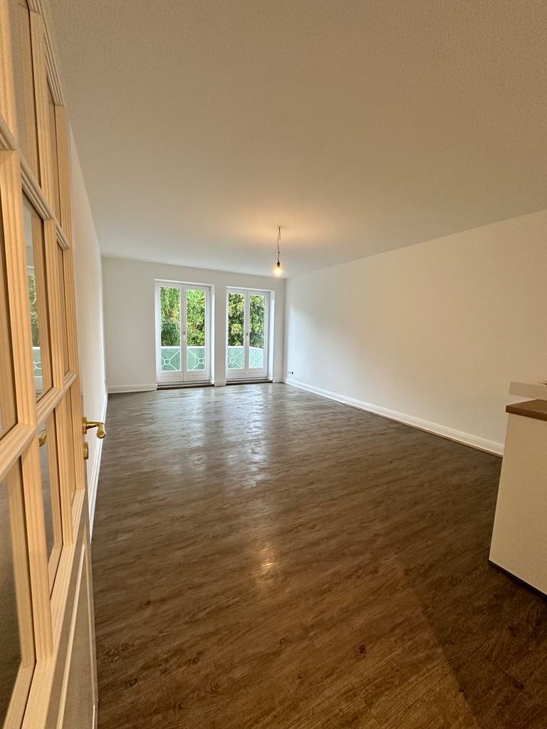 Jasne, przestronne wnętrze po remoncie z nową podłogą imitującą drewno, świeżo pomalowanymi ścianami i otwartymi, przeszklonymi drzwiami balkonowymi z widokiem na zieleń.