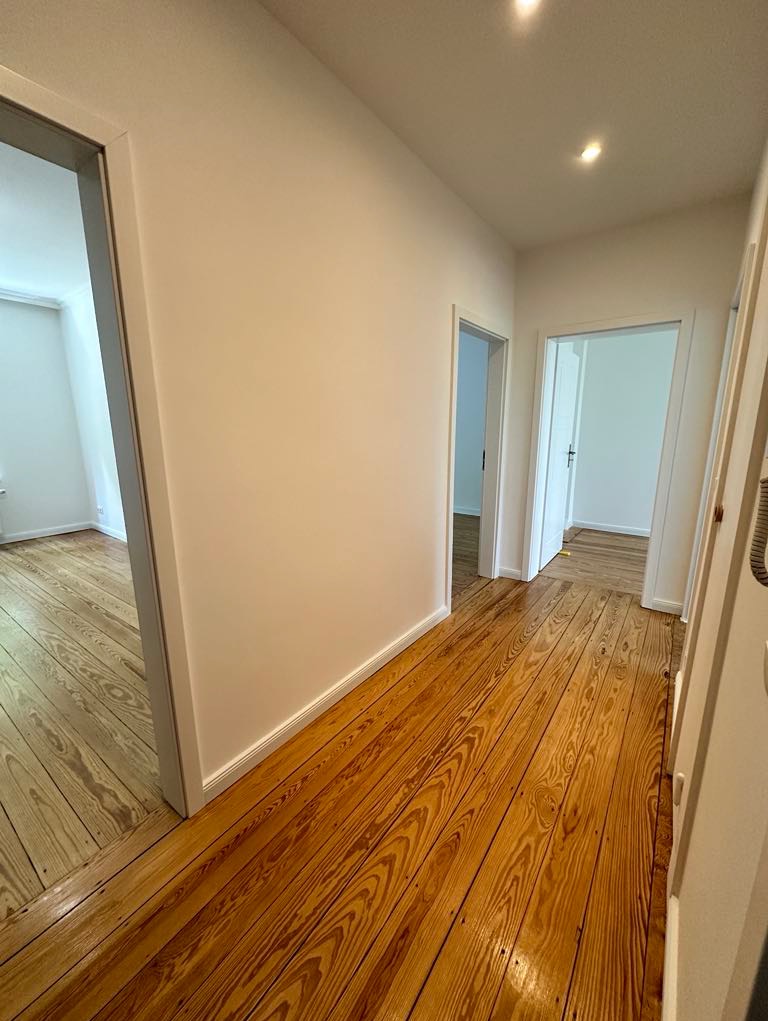 Korytarz po remoncie z odnowioną, lakierowaną podłogą z desek w odcieniach brązu i beżu, widoczne trzy pary drzwi do innych pomieszczeń, ściany pomalowane na biało.