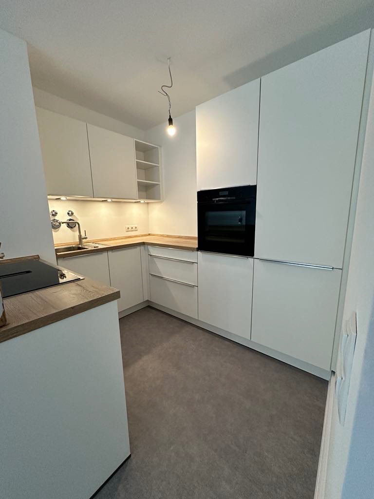 Nowoczesna, minimalistyczna kuchnia z białymi szafkami, drewnianym blatem i szarą podłogą. Widoczny zlew z baterią, płyta indukcyjna oraz piekarnik w zabudowie. Oświetlenie punktowe pod szafkami...