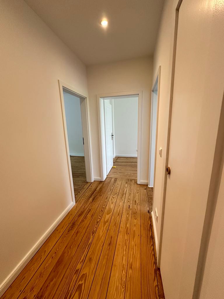 Korytarz w domu z odnowioną drewnianą podłogą, widoczne trzy wejścia do pomieszczeń z białymi ościeżnicami i białymi ścianami.