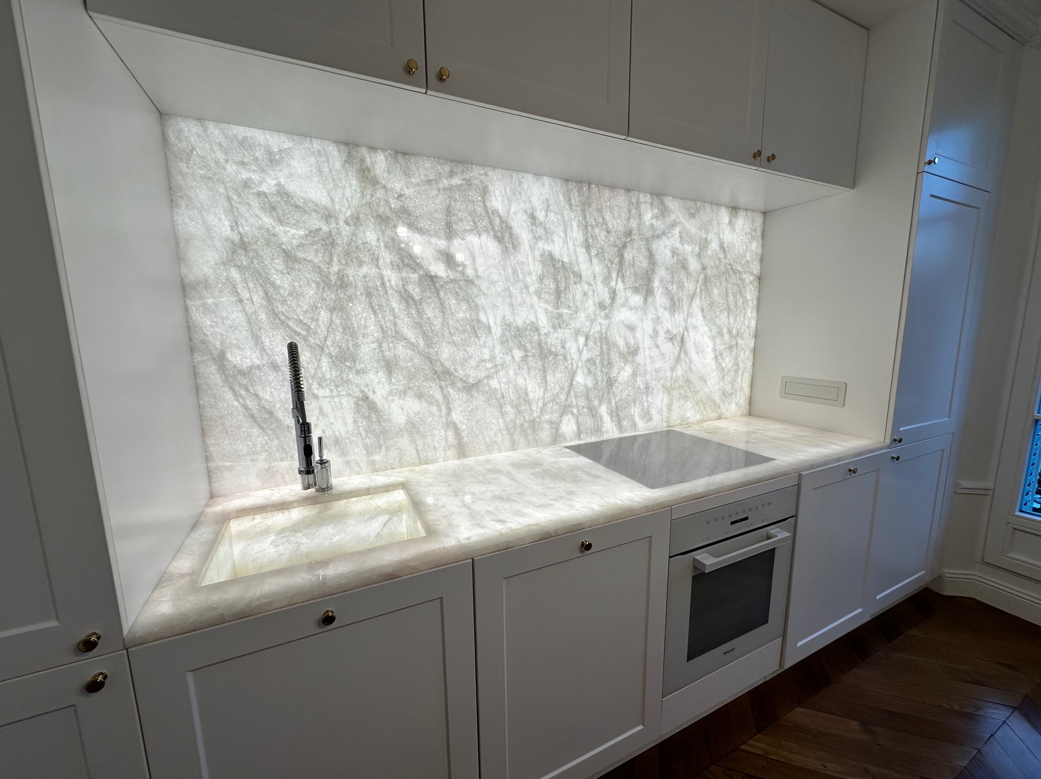 Biała kuchnia z podświetlanym blatem i panelem ściennym imitującym marmur, zintegrowany zlew i płyta indukcyjna, białe szafki z złotymi gałkami, parkiet w jodełkę.