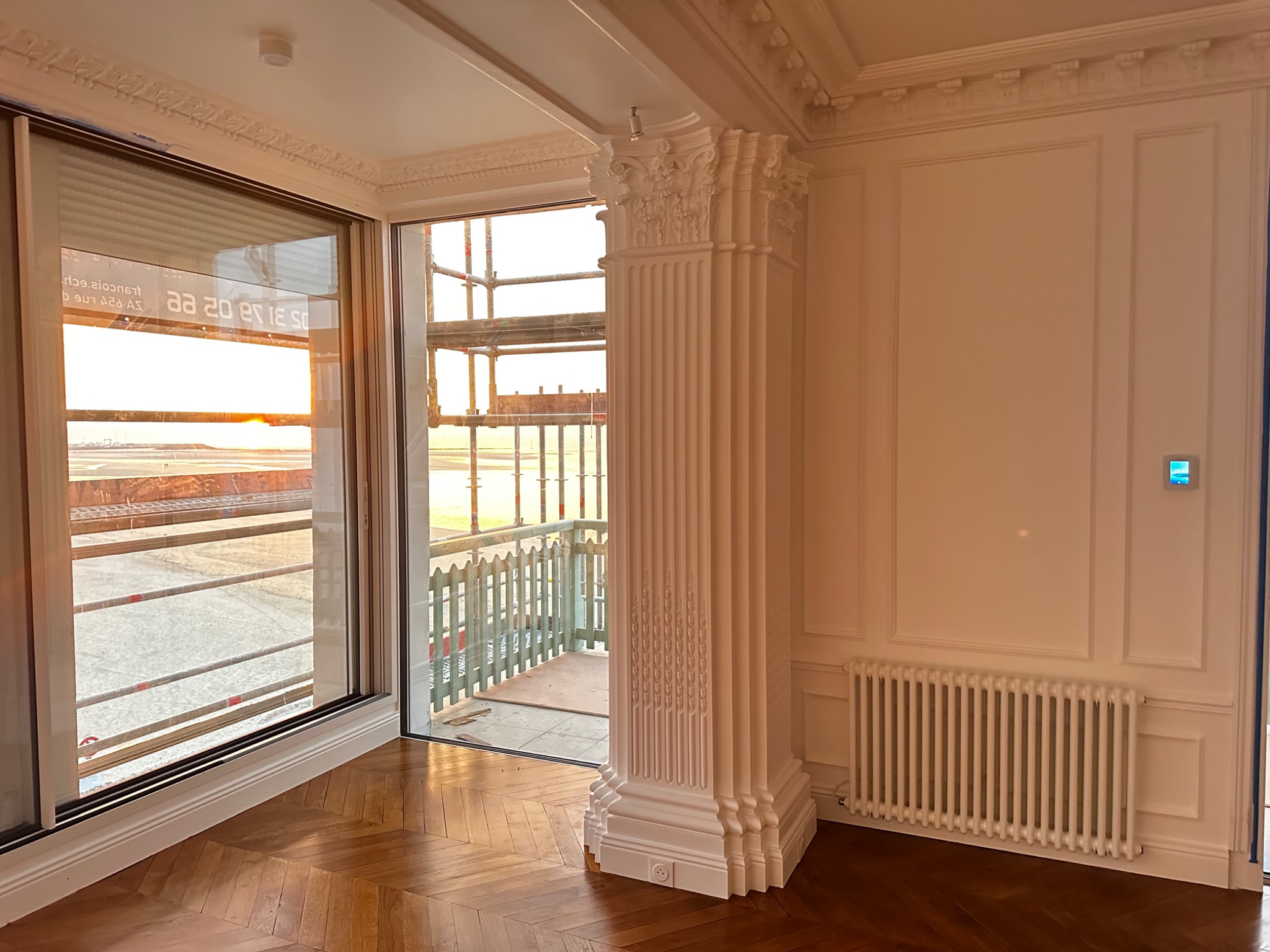 Wnętrze pokoju z parkietem ułożonym w jodełkę, ozdobną kolumną i listwami na ścianach, widok na rusztowania przez okno.