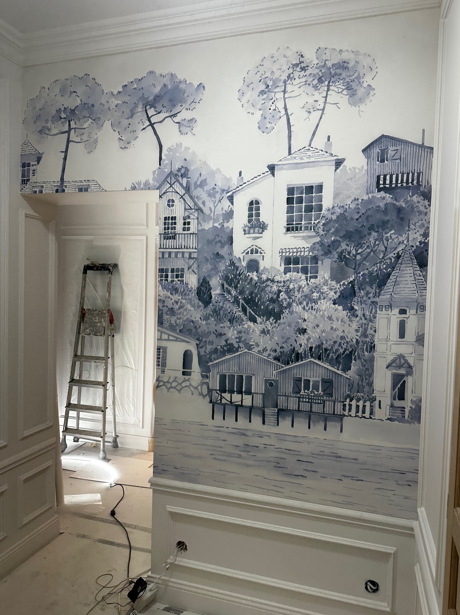 Malowidło ścienne w odcieniach błękitu przedstawiające nadmorski pejzaż z domami i drzewami, częściowo zakryte przez drabinę i zabezpieczenia malarskie w trakcie prac wykończeniowych.