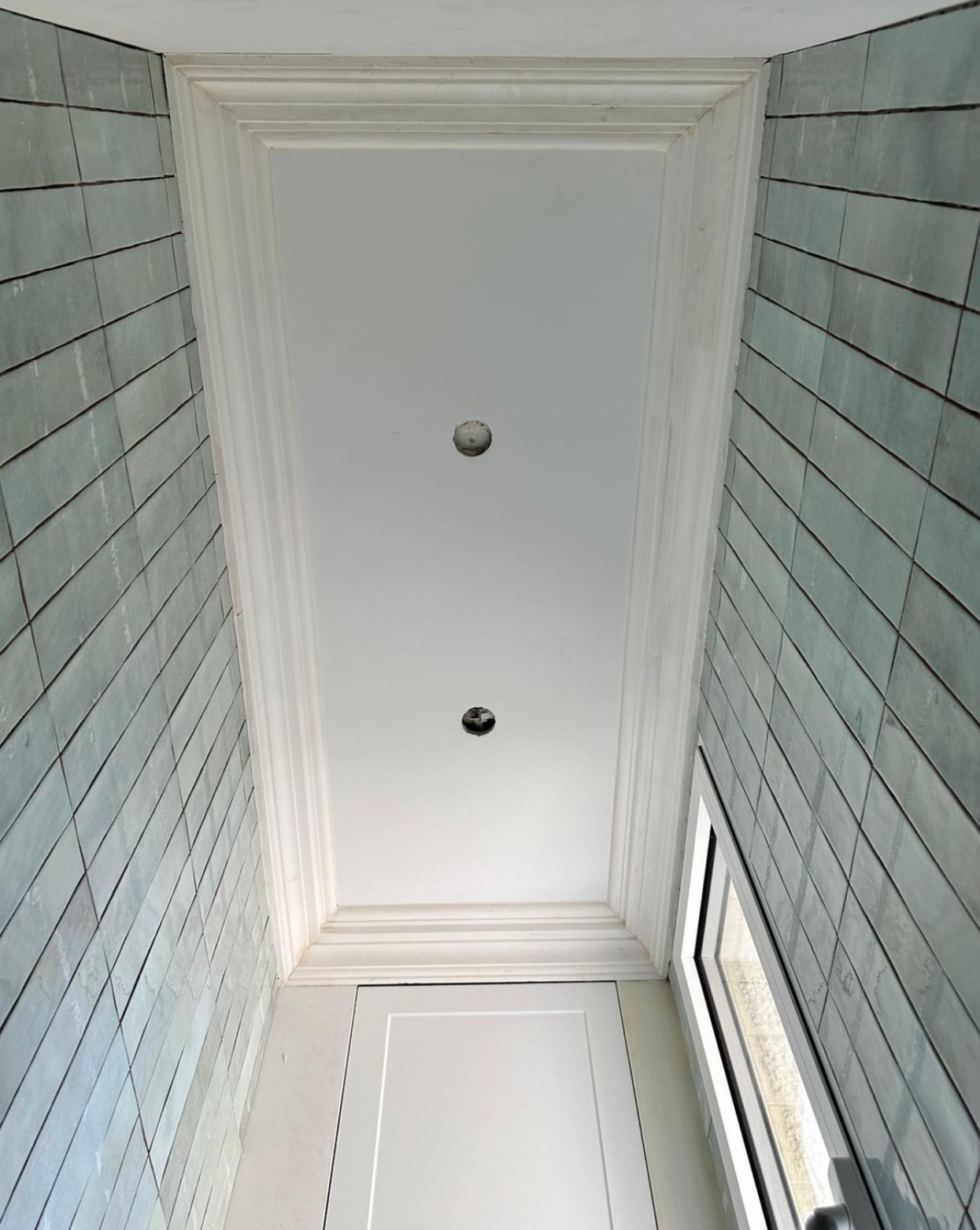 Wnętrze łazienki w trakcie wykańczania: ściany wyłożone ukośnie ułożonymi, zielonkawymi płytkami, biały sufit z ozdobną sztukaterią i dwoma otworami pod oświetlenie, widoczna górna część białych...