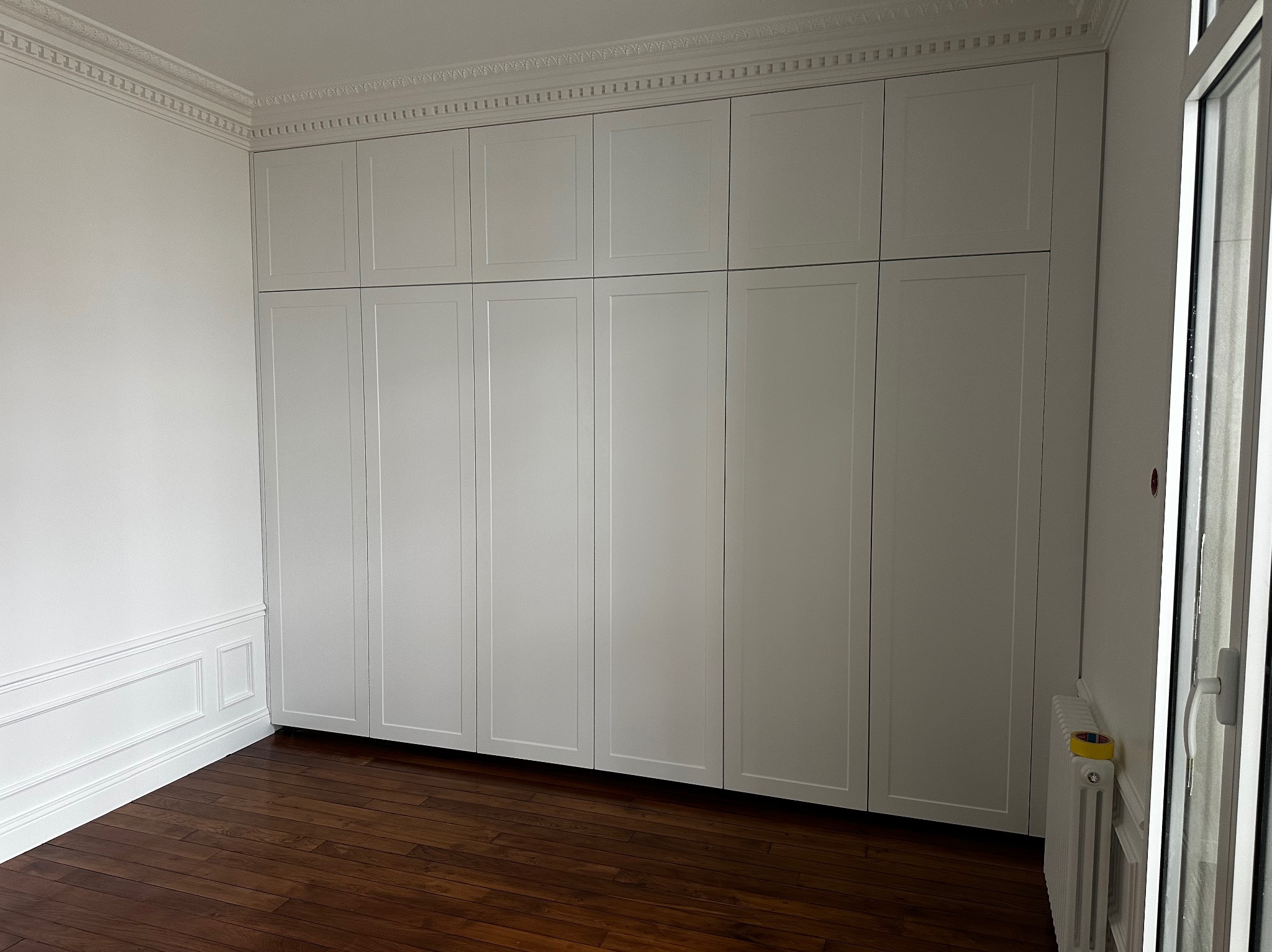 Biała zabudowa szafy wnękowej z frezowanymi frontami, widoczna listwa dekoracyjna pod sufitem i boazeria na ścianie, parkiet w ciemnym odcieniu brązu.