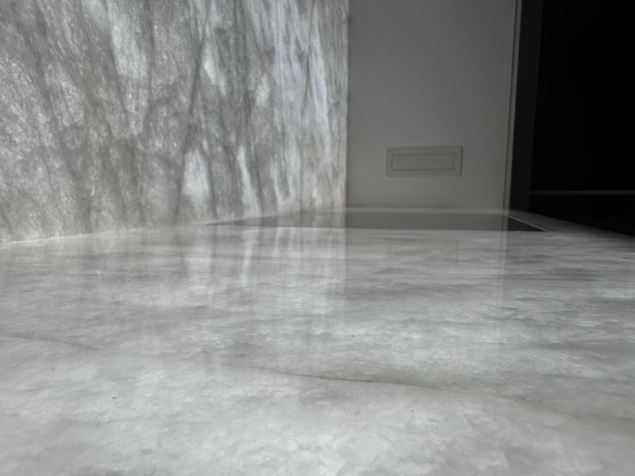 Błyszcząca, jasnoszara posadzka żywiczna w minimalistycznym wnętrzu, z dekoracyjną ścianą o fakturze przypominającej marmur i fragmentem białej ściany z gniazdkiem elektrycznym w tle.