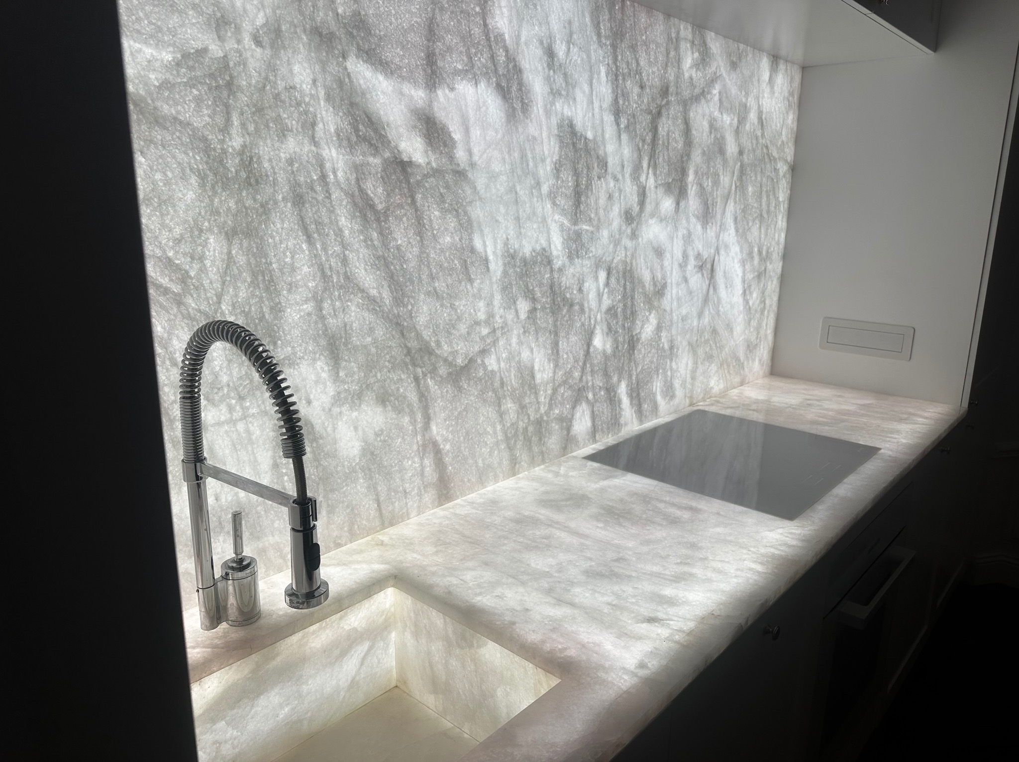 Kuchenny blat i ściana z podświetlanego, półprzezroczystego kamienia imitującego marmur, z nowoczesną baterią zlewozmywakową i płytą indukcyjną.