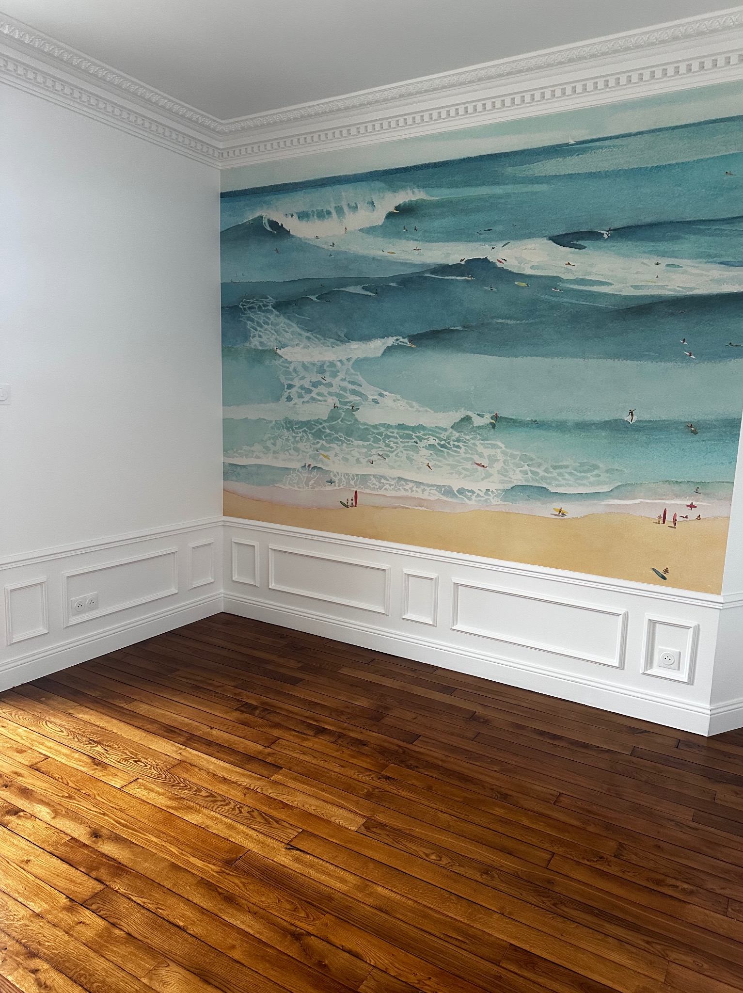 Pusty pokój z błyszczącą, brązową podłogą z desek, białe ściany z panelami dolnymi i górnym ozdobnym gzymsem, na jednej ścianie fototapeta z motywem plaży i fal morskich.